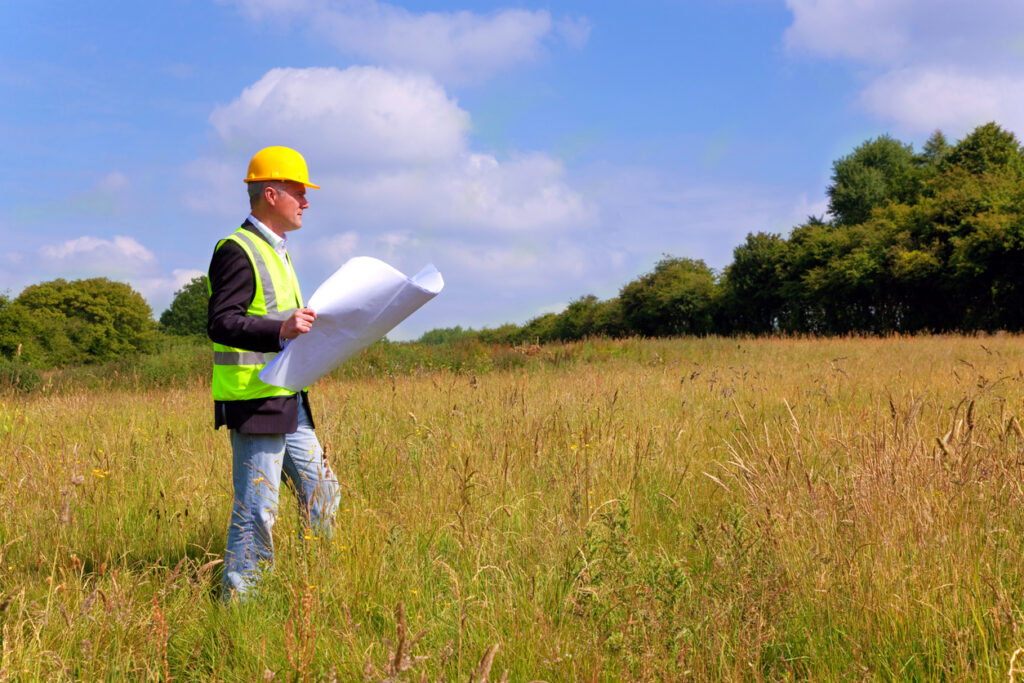 Phase I ESA specialist wearing site safety gear and holding a set of plans whilst surveying a new tower construction plot